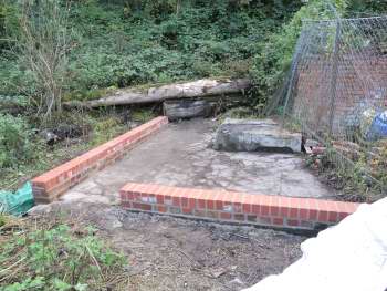 Low red brick wall runs around two edges of a grey concrete slab.   There is vegetation growing at the back of the slab