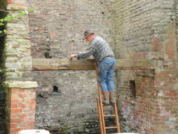A man uses a ladder to work on the top of a wooden beam stretched between two stone walls.