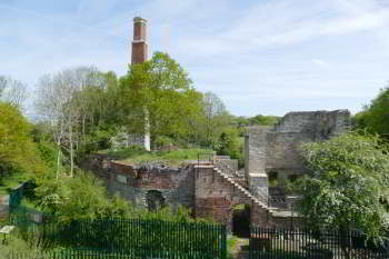 The Old Pit on a sunny spring day, with the chimney partly hidden by the leaves of the oak tree on the top of the heapstead and a Hawthorn in bloom partially hiding the Cornish Engine House.