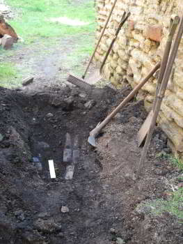 In the left foreground there are a number of objects lying in a hole dug into a path.  There are a number of tools leaning against a stone wall on the right.